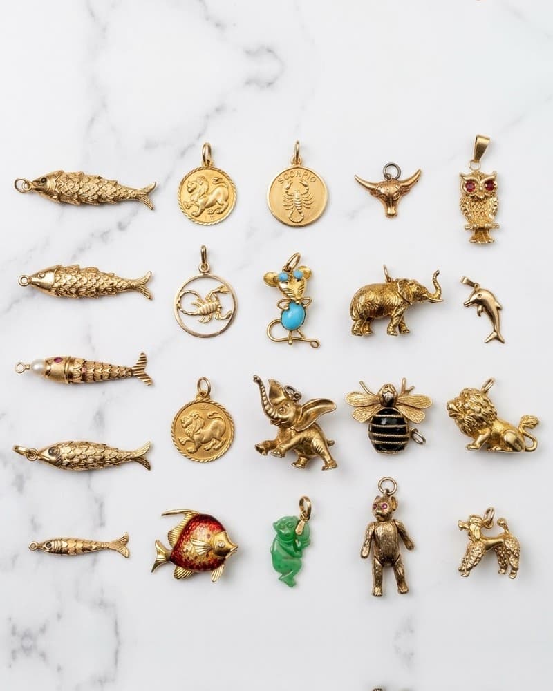 Twenty gold charms laid out on white marble — five articulated fish, a lion, an owl, a teddy bear, a turquoise mouse, an enamel fish, a jade monkey, a poodle.